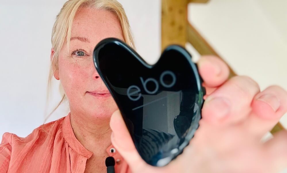 ebo founder, Kirstie Garrett, pictured in a coral shirt in front of a white background with light wooden beams holding a black obsidian The Wrinkle Eater TM face tool towards the camera, partially covering her face.